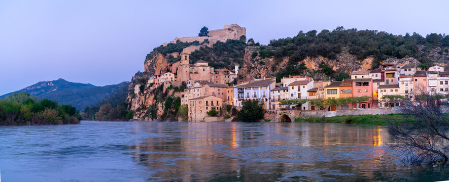 Miravet, Spain, a riverside village with a castle on a hill at dusk.