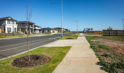 A newly developed suburban street lined with modern homes, freshly laid sidewalks, young trees supported by mulch beds, and a vacant lot awaiting further construction in Australia. © Doublelee