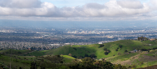A panoramic view of a city from rolling green hills under a cloudy sky.