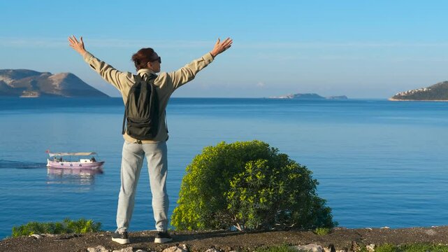Enjoying freedom with open arms by the sea. Female backpacker standing on rocky coastal viewpoint, raising arms while savoring expansive seascape, symbolizing freedom and wanderlust
