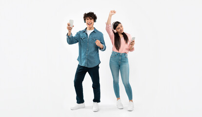 Fototapeta premium Digital Party. Happy Young Couple Listening To Music On Phones, Dancing And Singing Enjoying Musical Playlist Online Posing Over White Studio Background, Wearing Wireless Headphones. Panorama