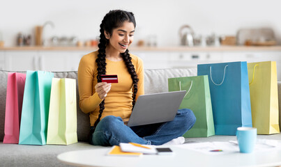 Happy Indian Female Making Online Shopping With Laptop And Credit Card At Home, Excited Eastern Woman Sitting On Couch Among Bright Shopper Bags, Enjoying Doing Internet Purchases, Free Space
