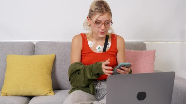 Young woman wearing holter monitor using laptop and smartphone on sofa