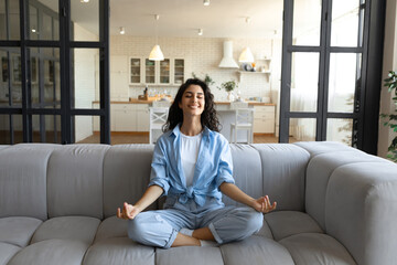 Obraz premium Stress management concept. Peaceful young lady in casual wear meditating with closed eyes on couch at home, full length. Calm millennial woman sitting on sofa in yoga pose, finding inner balance
