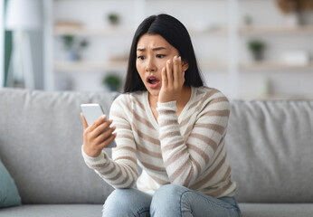 Obraz premium Oh No. Shocked Asian Woman Reading Bad Message On Smartphone While Sitting On Couch At Home, Confused Korean Female With Mobile Phone In Hands Emotionally Reacting To Unpleasant News, Free Space