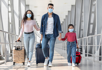 Obraz premium Travels During Pandemic. Arab Family Of Three Wearing Protective Medical Face Masks Walking With Suitcases At Airport Terminal, Arab Parents With Little Daughter Holding Hands While Going To Gate