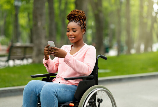 Happy handicapped black woman in wheelchair using smartphone, checking messages, browsing internet at city park. African American disabled lady with mobile device posting in social network outdoors - Powered by Adobe
