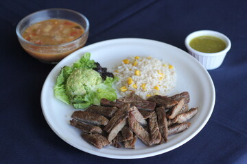 This vibrant plate showcases grilled steak, rice with corn, fresh guacamole, and a side of hearty soup, creating an enticing and satisfying meal experience.