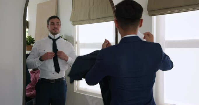 Groom tightening wedding-tie at living-room mirror groomsman draping jacket and dad adjusting lapel