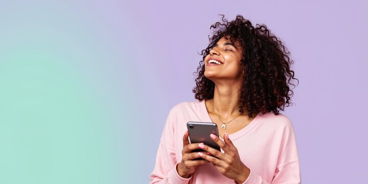 Happy woman using app joy, young black female joyful lively Adult human use smart phone, bold glow up face cute gaze. Modern studio light colors. Social mobile girl trendy style. Curly hair lady smile