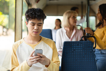 Portrait of young Asian guy traveling on public transit and using cellphone sitting on seat, reading sms message looking at mobile phone screen, chatting online during trip ride inside crowded vehicle