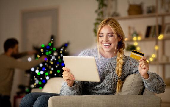 Happy woman shopping online on Christmas, using digital tablet and credit card, xmas sales concept, copy space. Smiling young lady with pad sitting at home and buying xmas gifts for her family