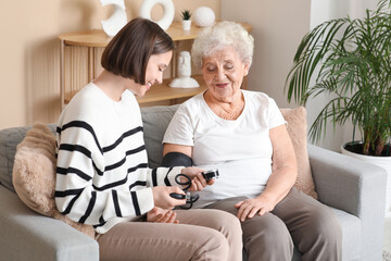 Young nurse with senior woman measuring blood pressure on sofa at home