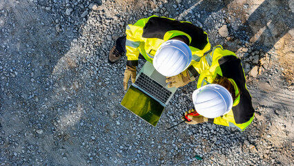 Aerial view of two construction workers in safety gear reviewing plans on a laptop amidst metal beams, emphasizing teamwork, precision, and the role of technology in modern construction projects.