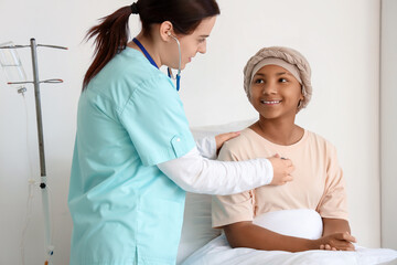 Obraz premium Female doctor with stethoscope listening to African-American girl after chemotherapy in hospital room