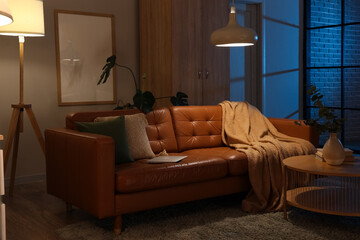 Stylish interior of living room with cozy sofa, coffee table and glowing lamps at night