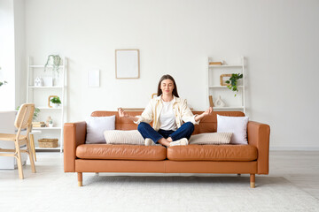 Beautiful young woman meditating on brown couch at home