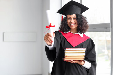 Happy African-American female graduate with diploma and books near window