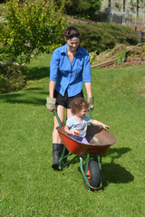 Young adult woman carry her toddler inside a wheelbarrow around home garden