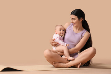 Sporty young woman with her little baby sitting on yoga mat against beige background