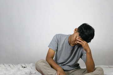 Portrait Of Depressed Young Indonesian Man Sitting On Bed At Home, Suffering Life Problems Or Mental Breakdown, Feeling Despair, Copy Space
