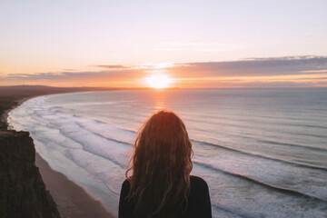 Fototapeta premium A serene photograph of a person with long brown hair, viewed from behind, sitting on a rocky cliff edge overlooking a vast ocean at sunset. 