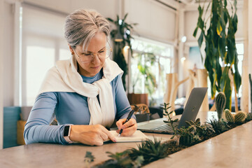 Focused senior woman with gray hair writing in notebook while working on laptop in cozy modern cafe
