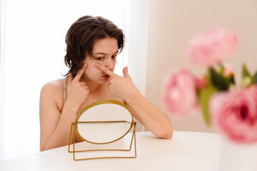 Stressed sad brunette young woman wearing beige top looking at mirror and touching her face having...