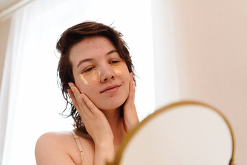 Stressed sad brunette young woman wearing beige top looking at mirror and touching her face having...