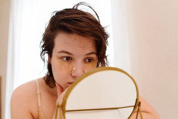Stressed sad brunette young woman wearing beige top looking at mirror and touching her face having...