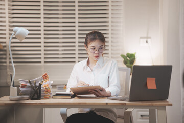 Asian businesswoman working late at night in office, reviewing documents under desk lamp, showing...