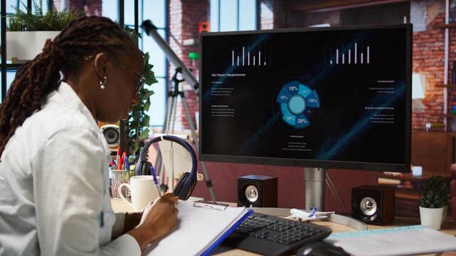 Remote worker at home watching business conference on PC monitor, looking over statistics on documents. Freelancer in apartment looking at presentation and checking paperwork files, camera A