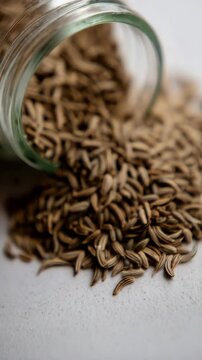 Close-up shot of cumin seeds spilling from a clear glass jar onto a white surface, showcasing the spice's texture and color with shallow depth of field.