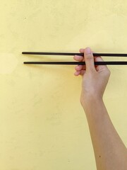 The image shows a right hand holding a pair of black chopsticks againts a light yellow wall