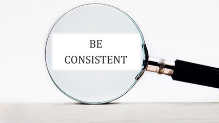 Business concept text BE CONSISTENT focused through a magnifying glass