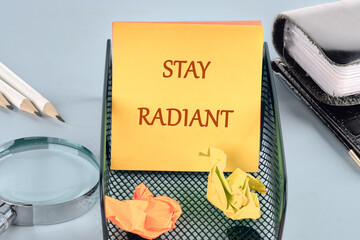 A bright yellow sticky note with the message stay radiant sits in a black mesh organizer at a desk. Crumpled pieces of paper and office tools create a busy yet inspiring workspace environment