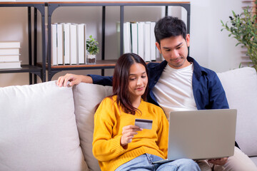 Fototapeta premium Happy asian couple using laptop and credit card for online shopping together in living room at home, man and woman smiling and relax with enjoy a digital lifestyle, convenient payment.
