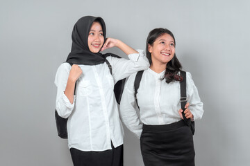 Two cheerful Asian teenage girls wearing school uniforms and backpacks, smiling and looking away, isolated on gray background. Concept of back to school season.