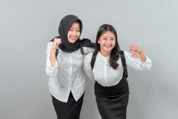 Two Asian schoolgirls smiling and posing energetically with backpacks on their shoulders. Confident back to school spirit, showing teamwork and enthusiasm. Isolated on gray background.