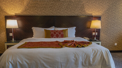 The double bed is covered with white sheets. The red bedspread is neatly and beautifully laid. Pillows at the headboard. The bedside lamps are lit. Golden beige patterned wallpaper on the wall.  