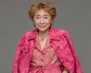 Elegant elderly woman smiling confidently wearing a pink jacket and patterned blouse with short curly hair and jewelry in front of a gray background