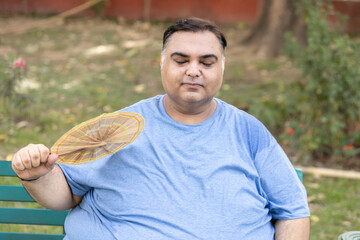 Overweight indian man using hand fan in hot summer day. Fat guy suffering from heat wave due to...