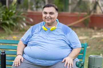 Happy Plus size indian man relaxing wearing portable cooling fan around neck get relief in hot...