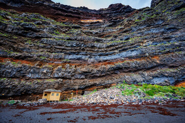 Small wooden structure nestled against dramatic layered volcanic cliffs and surrounded by black sand, illustrating scale, solitude and natural resilience.