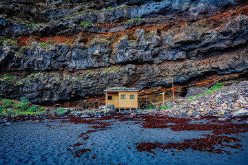 Small wooden structure nestled against dramatic layered volcanic cliffs and surrounded by black sand, illustrating scale, solitude and natural resilience.