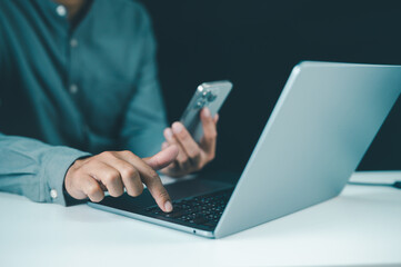 A person uses a smartphone and laptop simultaneously, indicating multitasking or remote work in a modern office setting.