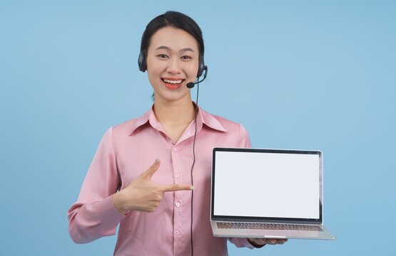 Smiling woman wearing headset points to blank laptop screen against blue background during online communication session