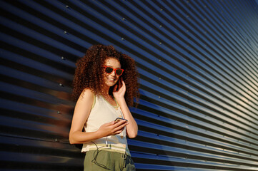 young woman using smartphone outdoors, smiling in sunlight, wearing casual tank top and sunglasses....