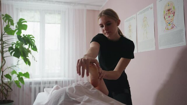 Massage therapist wearing black lifting client's foot for therapeutic massage in tranquil spa room, featuring bright window light, indoor plant, and wall charts illustrating human anatomy