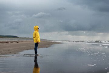 Boy in yellow jacket standing by stormy sea, looking at sea.